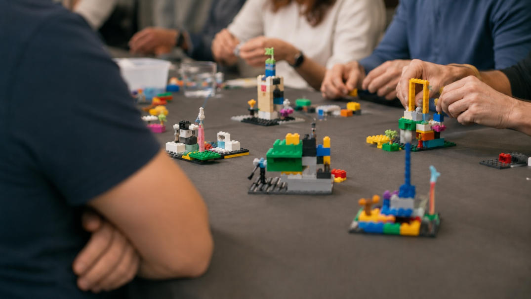 Participants concentrate on building LEGO SERIOUS PLAY models while one person in the foreground still watches with crossed arms